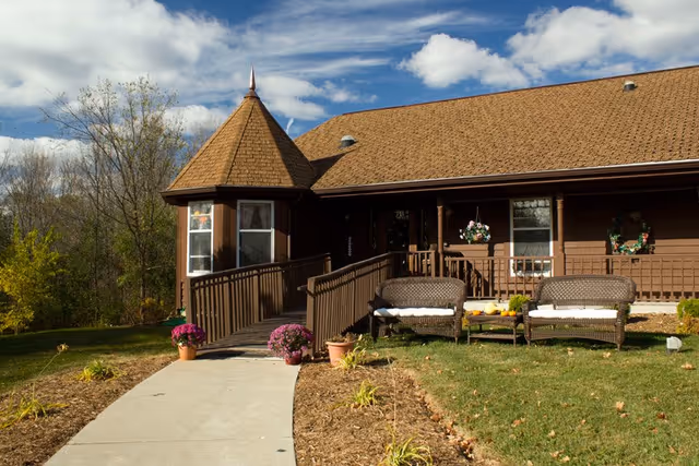 Exterior view of a brown Victorian-style senior living home with a pointed turret, a ramp leading to the entrance, and two wicker chairs with cushions on a grassy lawn. There are potted flowers near the ramp and hanging flower baskets on the porch under a partly cloudy sky.
