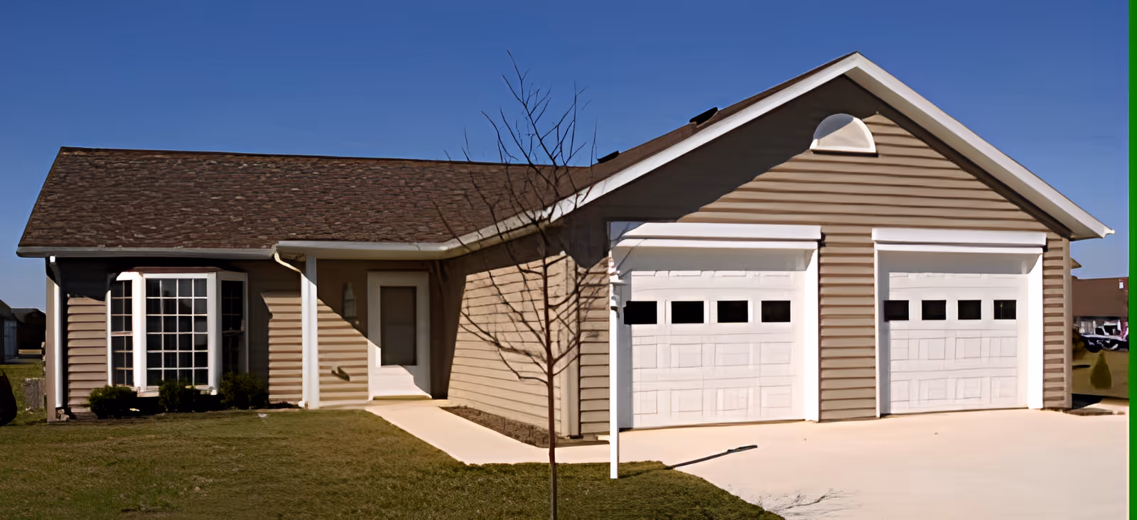 Exterior view of a single-story building with beige siding, a brown shingled roof, a white front door, a bay window with multiple panes, and a double garage with white doors. There is a small tree without leaves in the front yard and a clear blue sky in the background.