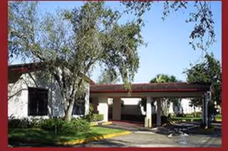 Exterior view of a single-story building with a covered entrance driveway, surrounded by trees and greenery under a clear blue sky.