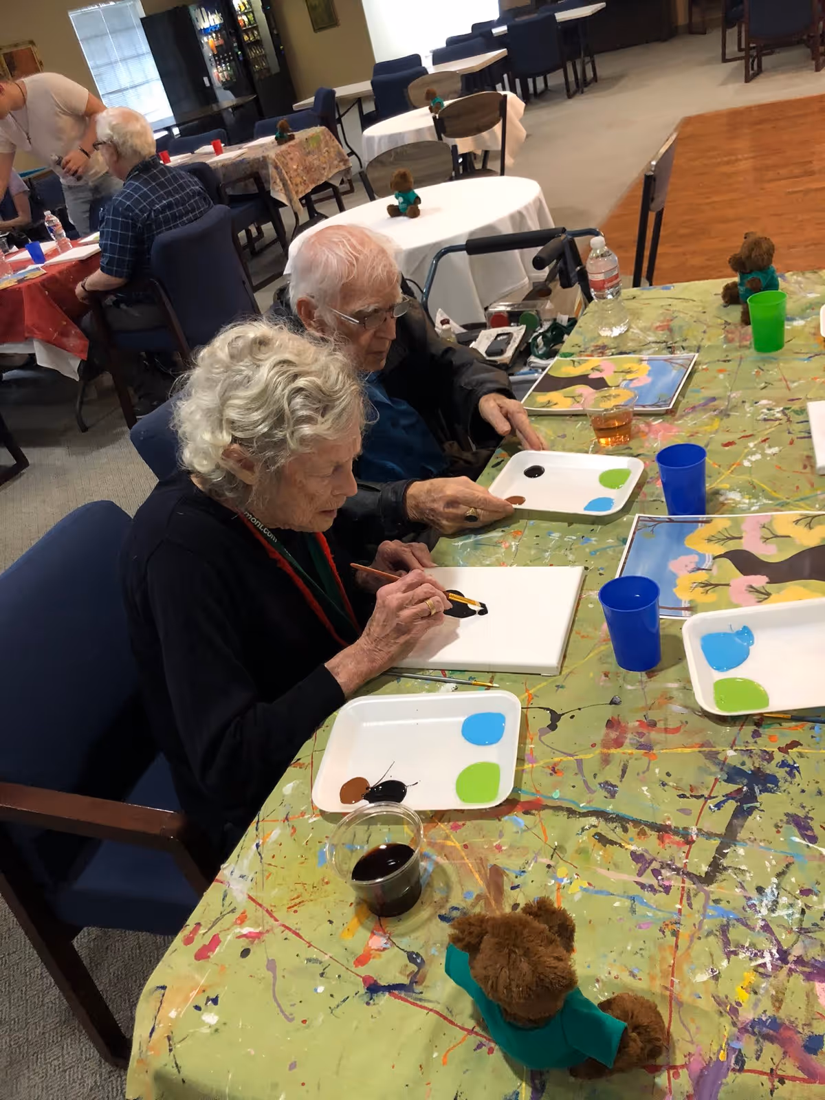 Two elderly individuals seated at a paint-splattered table engaged in a painting activity. The woman in the foreground is painting on a canvas with black and yellow paint, while the man beside her holds a paint palette. The table has paint trays with various colors, cups, and small teddy bears wearing green shirts. In the background, other elderly people are seated at tables covered with tablecloths in a communal room with chairs and a vending machine.