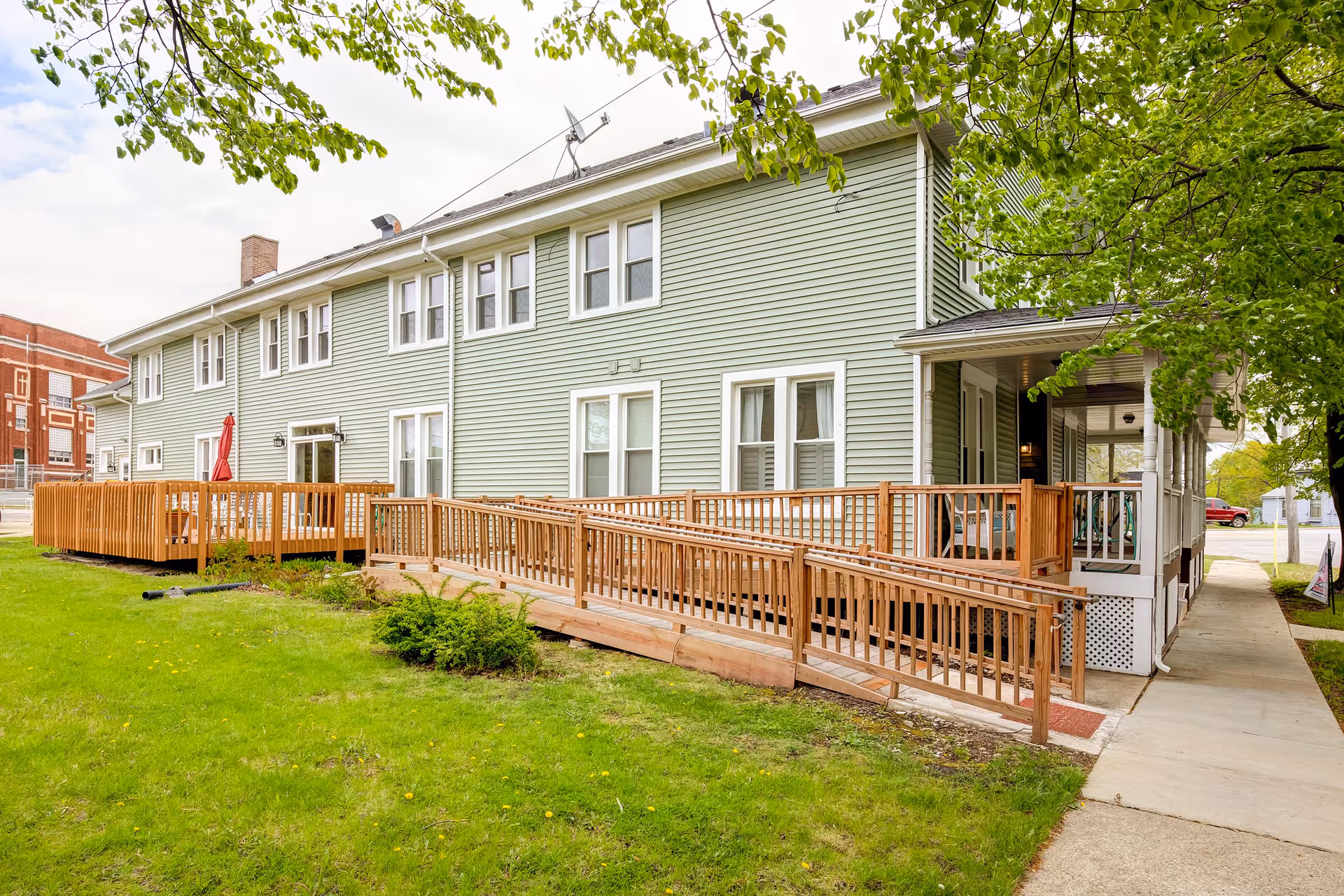Exterior view of a two-story light green senior living facility building with white trim, featuring a wooden wheelchair ramp and a small porch area. The building is surrounded by green grass and trees, with a sidewalk running alongside it.