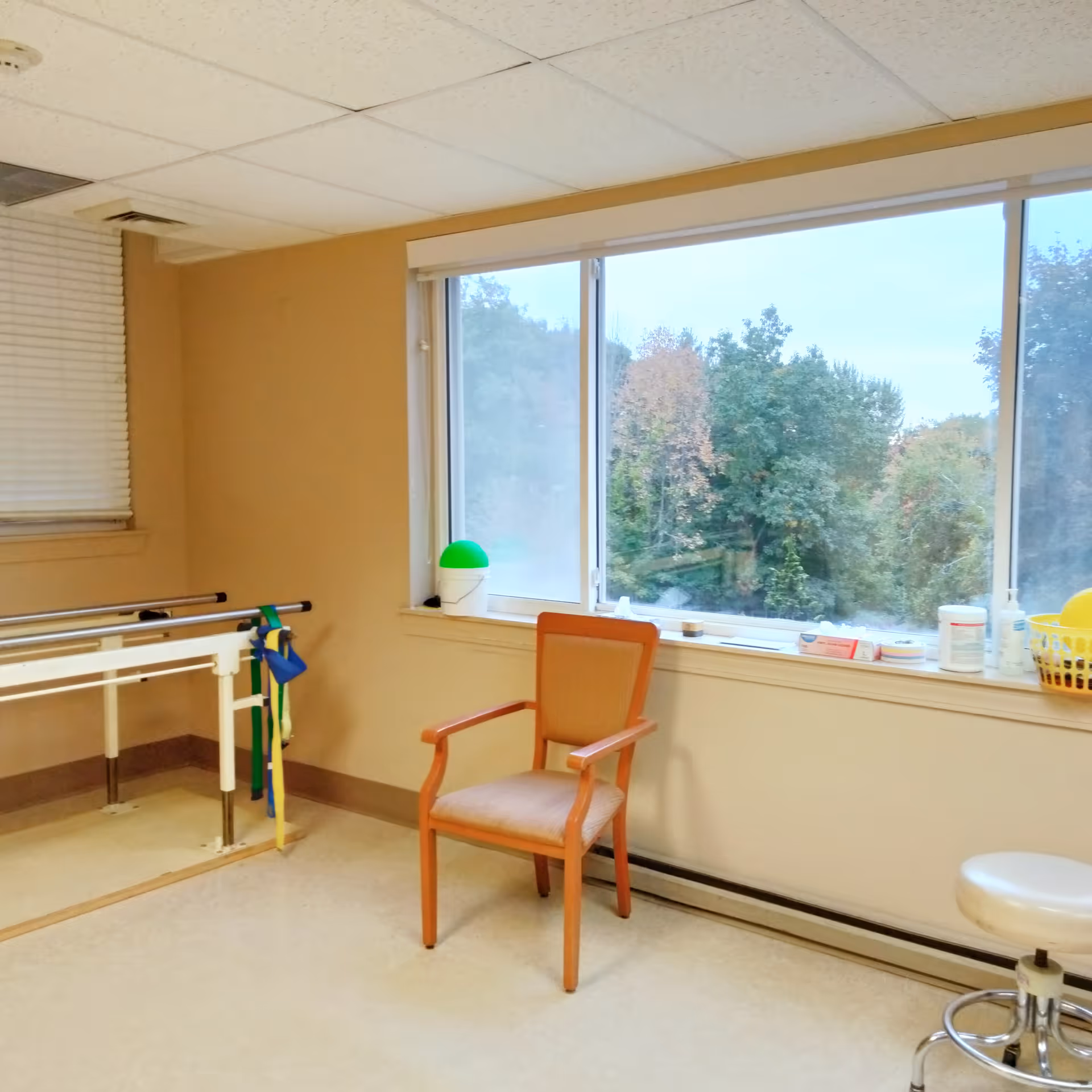 A rehabilitation room with a wooden chair, a white stool on wheels, parallel bars for walking exercises, and a large window showing trees outside. Various medical supplies are placed on the windowsill.