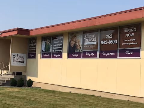 Exterior view of a single-story building with beige walls and a red roof trim. Several large windows display signs for Home Instead Senior Care, listing services such as Alzheimer's care, assistance care, and hospice support. The signs also mention hiring for CNA or HCA/C positions with no experience required. There is a small set of stairs leading to a door on the left side of the building, and a grassy area in front.