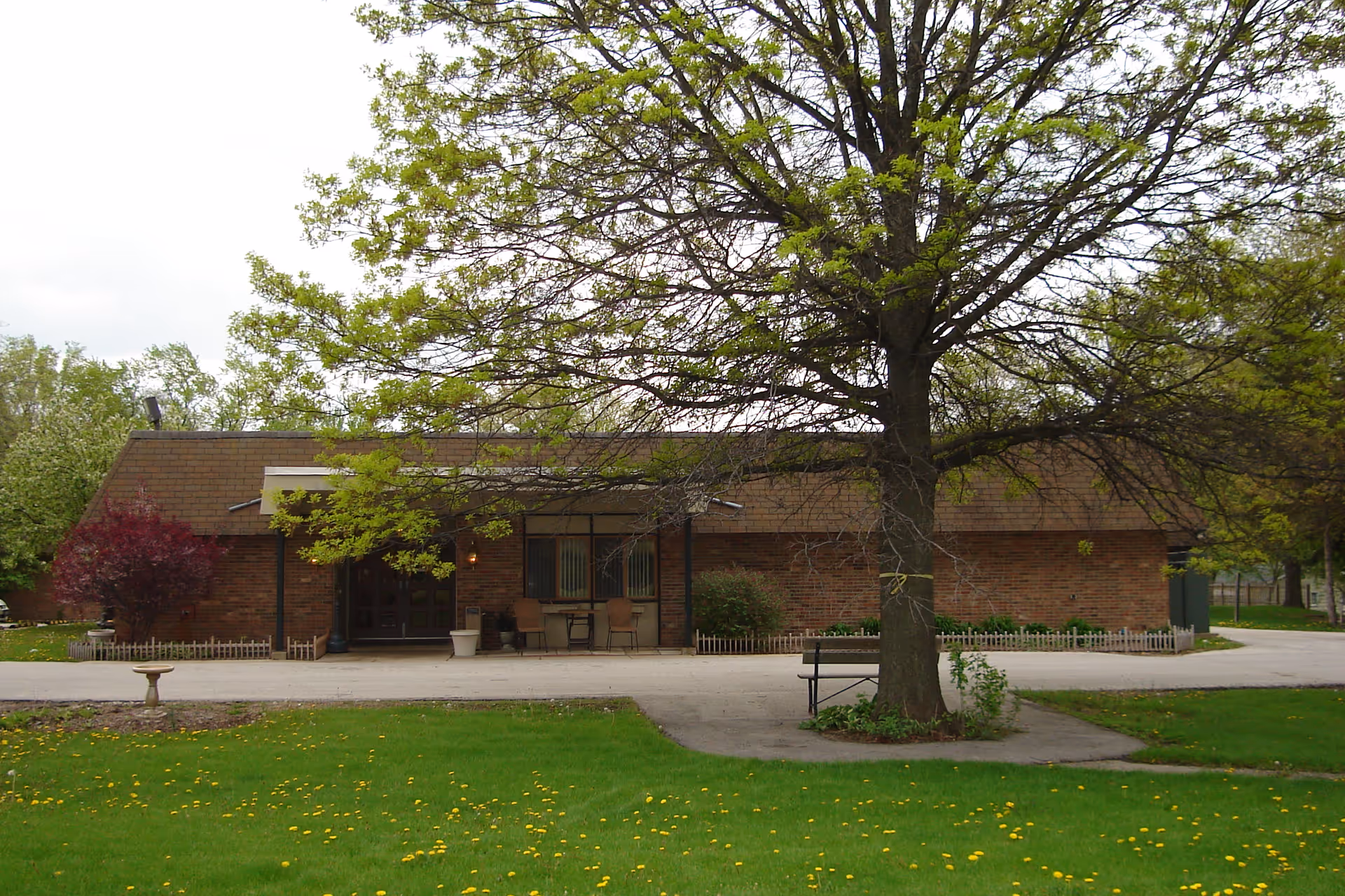 Exterior view of a single-story brick building with a sloped roof, surrounded by green grass and trees. There is a large tree in the foreground with a bench underneath it, and a small paved area leading to the building entrance with two chairs and a table. The sky is overcast.