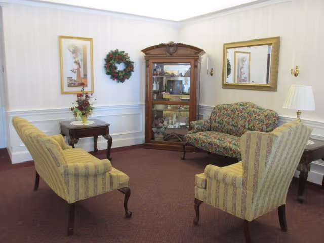 A cozy sitting area in a retirement community featuring two striped armchairs and a floral patterned loveseat arranged around a small wooden side table with a floral arrangement. A wooden display cabinet with glass doors stands against the wall, flanked by a framed picture and a decorative wreath. A large mirror hangs on the adjacent wall above another side table with a lamp. The room has white walls with chair rail molding and a carpeted floor.