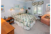 A cozy bedroom featuring a bed with floral patterned bedding, a wooden nightstand with a lamp and decorative items, a window with floral valance curtains, a peach-colored armchair, and framed artwork on the light blue walls.