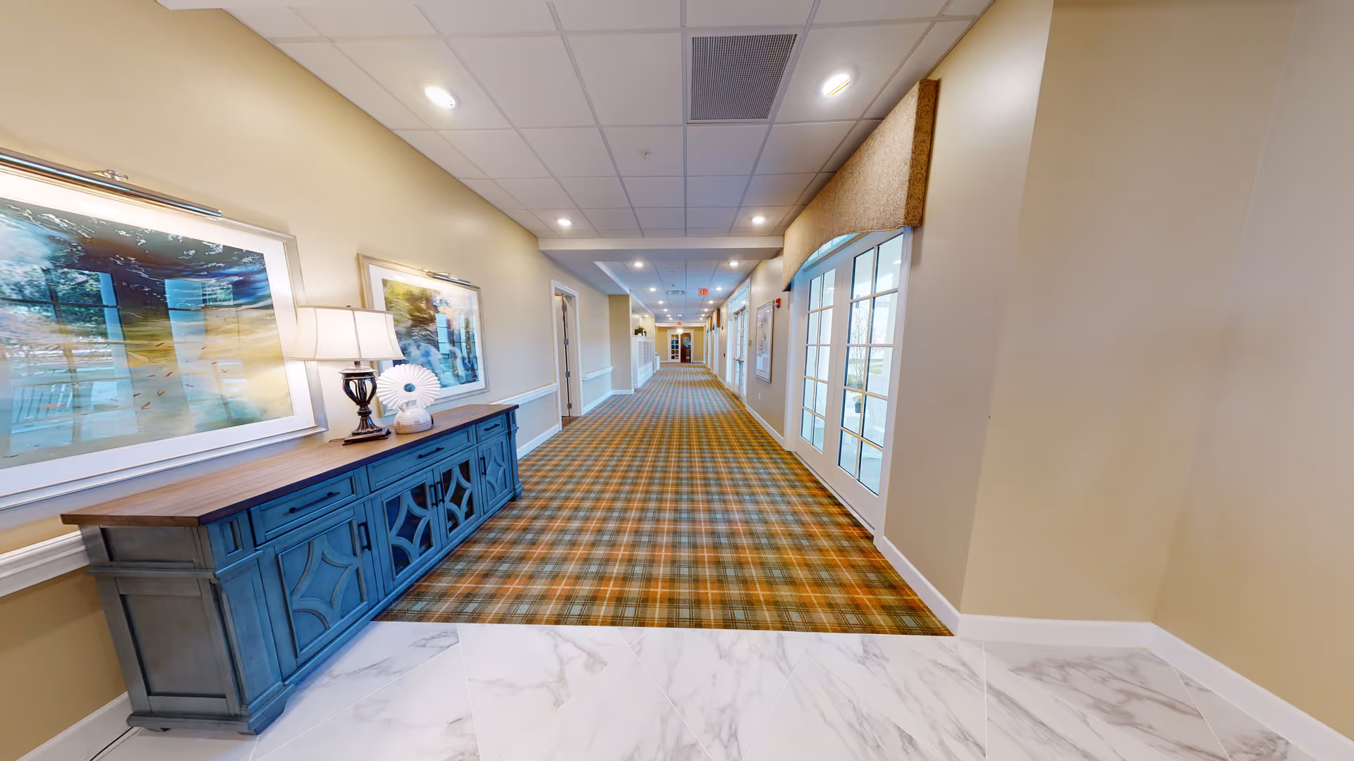 A long, well-lit hallway in a senior living facility with beige walls, a patterned carpet, and white marble tile flooring at the entrance. On the left side, there is a blue wooden console table with a lamp and decorative items, and framed artwork hanging above it. On the right side, there are glass doors with a valance above them, leading to an outdoor area.