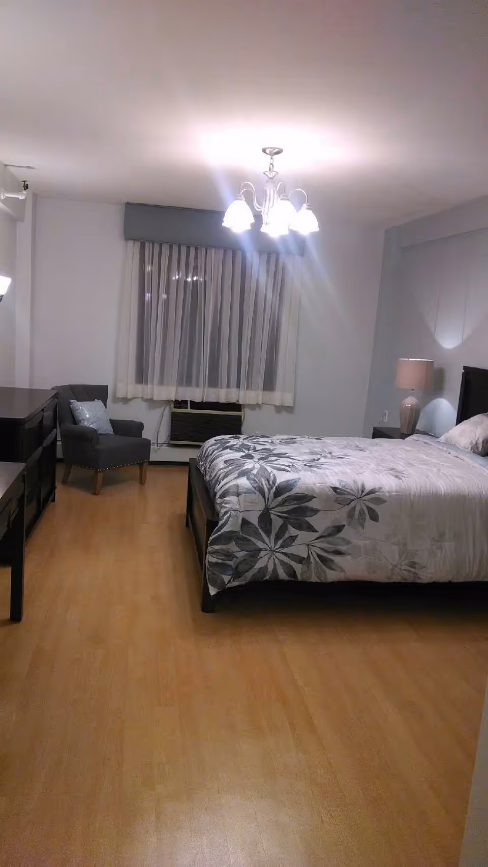 A bedroom with a bed covered in a floral patterned comforter, a nightstand with a lamp, a gray armchair with a pillow, a dresser, and a window with sheer curtains. The room has wooden flooring and a ceiling light fixture with multiple bulbs.