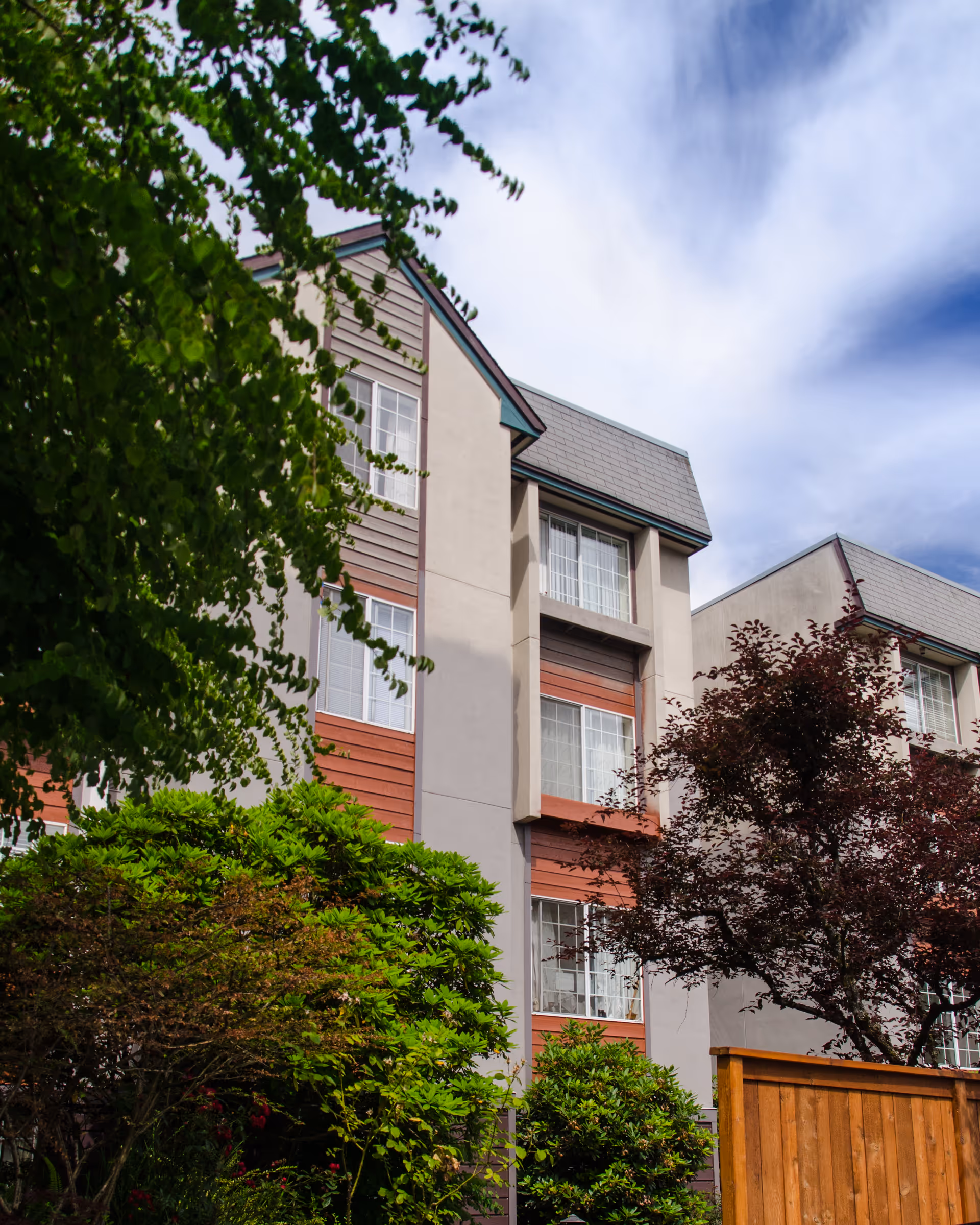 Exterior view of a multi-story residential building with beige and reddish-brown siding, surrounded by green trees and bushes under a partly cloudy sky.