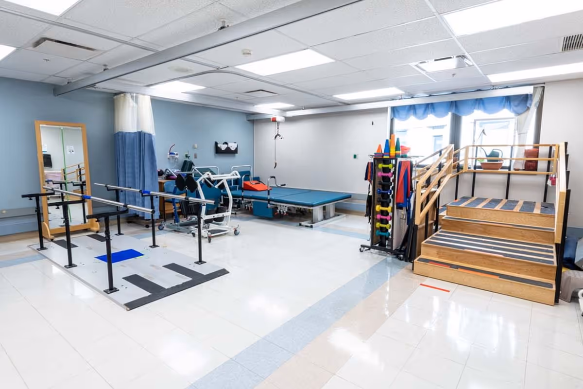 A physical therapy or rehabilitation room with parallel bars for walking exercises, a large mirror, a padded therapy table, exercise equipment including weights and cones, and a wooden staircase with handrails for mobility training. The room has tiled floors, light blue and white walls, and fluorescent ceiling lights.
