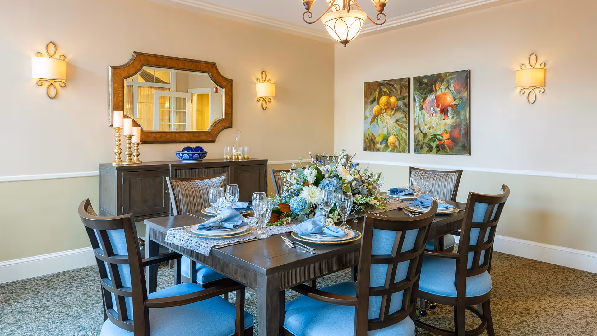 A well-lit dining room with a dark wooden table set for six people. The table is decorated with a floral centerpiece, blue napkins, glassware, and plates. Surrounding the table are six wooden chairs with blue cushions. On the wall behind the table, there are two colorful paintings of fruit and a large decorative mirror above a dark wooden sideboard. The room has beige walls with white trim, carpeted flooring, and elegant wall sconces and a chandelier providing warm lighting.