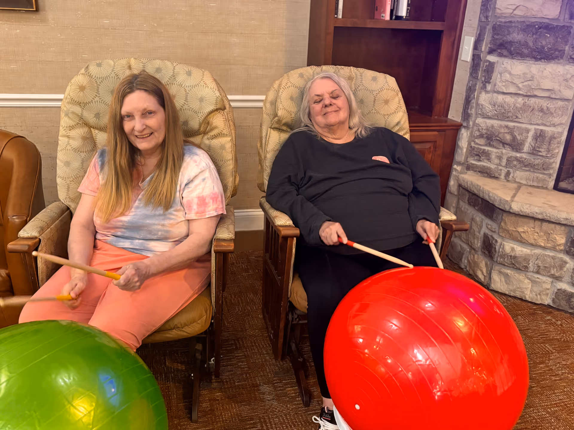 Two elderly women sitting in cushioned armchairs indoors, each holding drumsticks and playing on large exercise balls, one green and one red. They are in a cozy room with a stone fireplace and wooden shelves in the background.