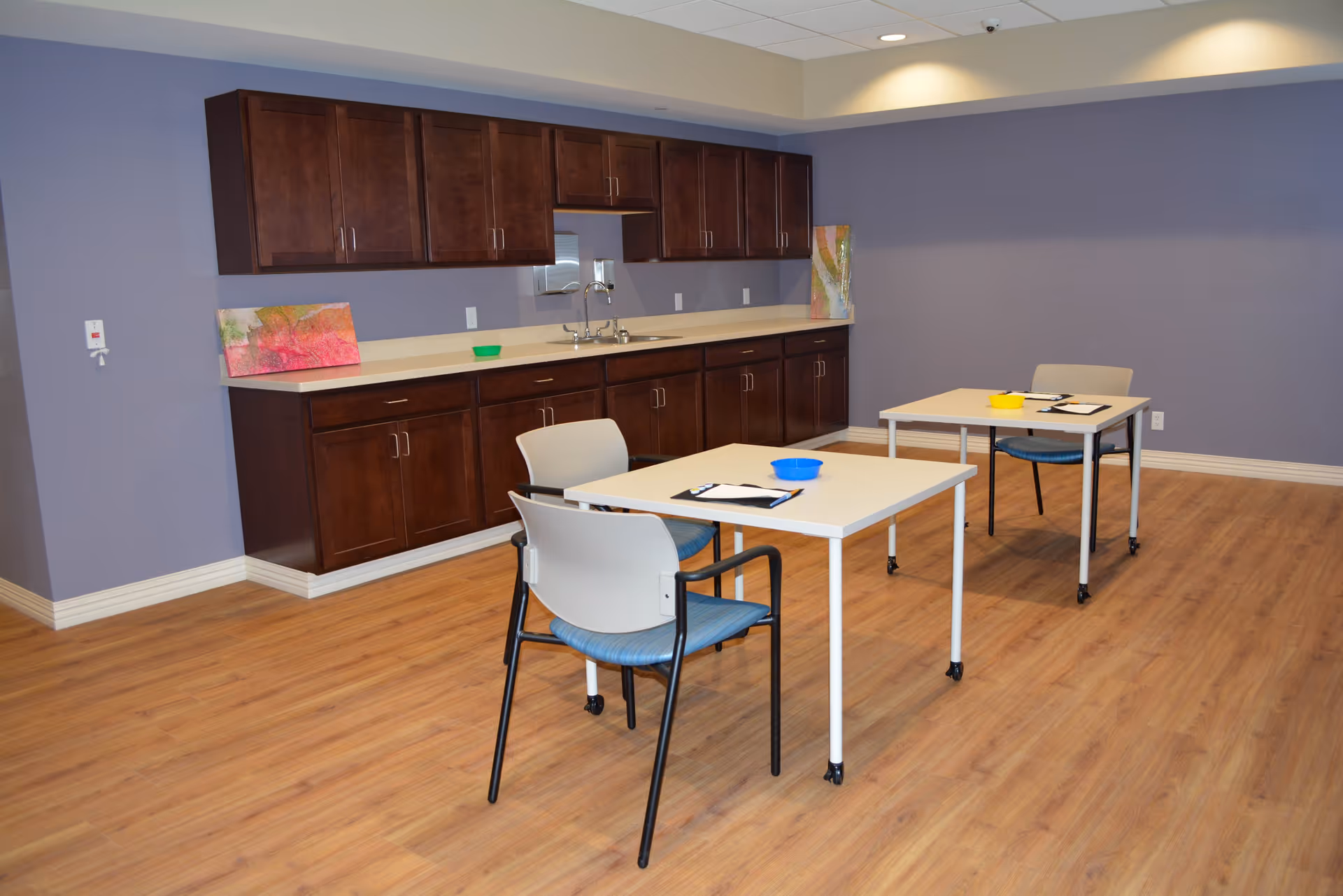 A room with light wooden flooring and lavender walls featuring a kitchenette with dark brown cabinets and a countertop with a sink. Two white tables with wheels are positioned in the room, each accompanied by a chair with a blue seat cushion. On the tables are clipboards and colorful bowls. Two pieces of abstract artwork lean against the wall on the countertop.