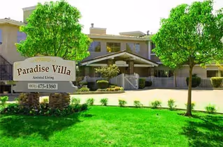 Exterior view of Paradise Villa Assisted Living facility showing a two-story building with a covered entrance, surrounded by green trees and a well-maintained lawn. A sign in front reads 'Paradise Villa Assisted Living' with a phone number.