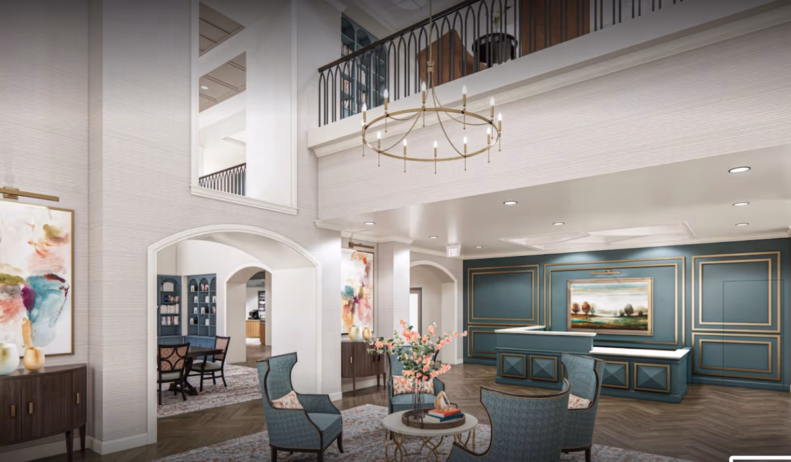 A spacious and elegant senior living facility lobby with high ceilings and a large circular chandelier. The room features a reception desk with teal and gold paneling, a round table with four upholstered chairs, floral centerpiece, and abstract artwork on the walls. There is a mezzanine level with a black railing and built-in bookshelves visible in the background.