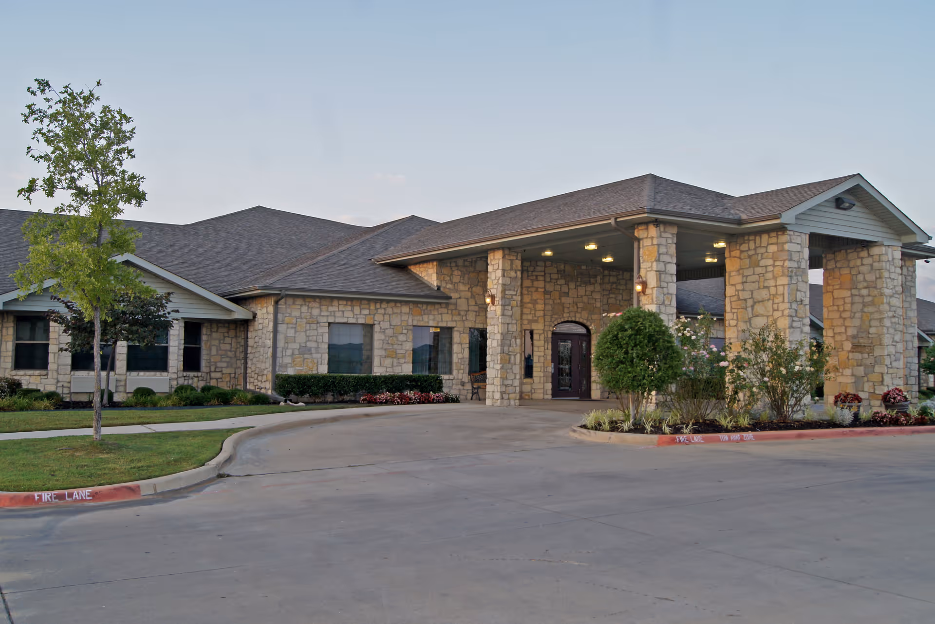 Exterior view of Crowley Nursing and Rehabilitation facility showing a stone building with a covered entrance supported by stone pillars, surrounded by landscaping and a driveway.