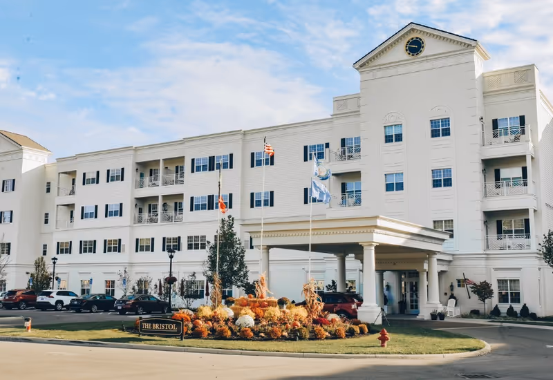 Exterior view of The Bristol Senior Living at Worthington Hills, a large white multi-story building with multiple windows and balconies. The entrance features a covered driveway with columns, and there are three flagpoles with flags in front of the building. A landscaped area with flowers and plants is visible near the entrance, along with parked cars in the parking lot.