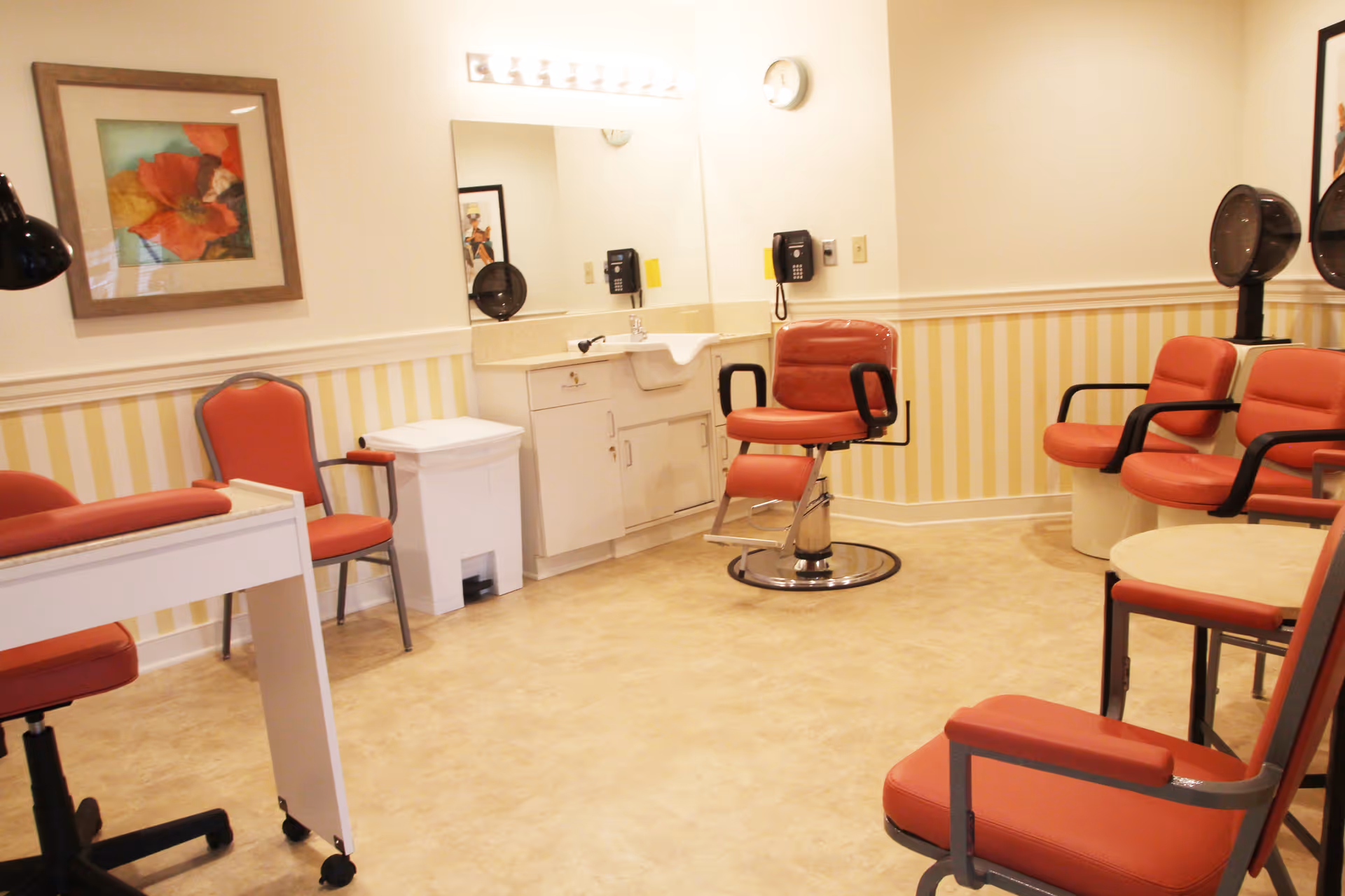 A bright room with several red cushioned chairs, including a salon-style chair in front of a sink and mirror. The walls have yellow and white vertical stripes with a framed floral picture and a clock. There are hair drying stations with hooded dryers and a small round table with chairs.