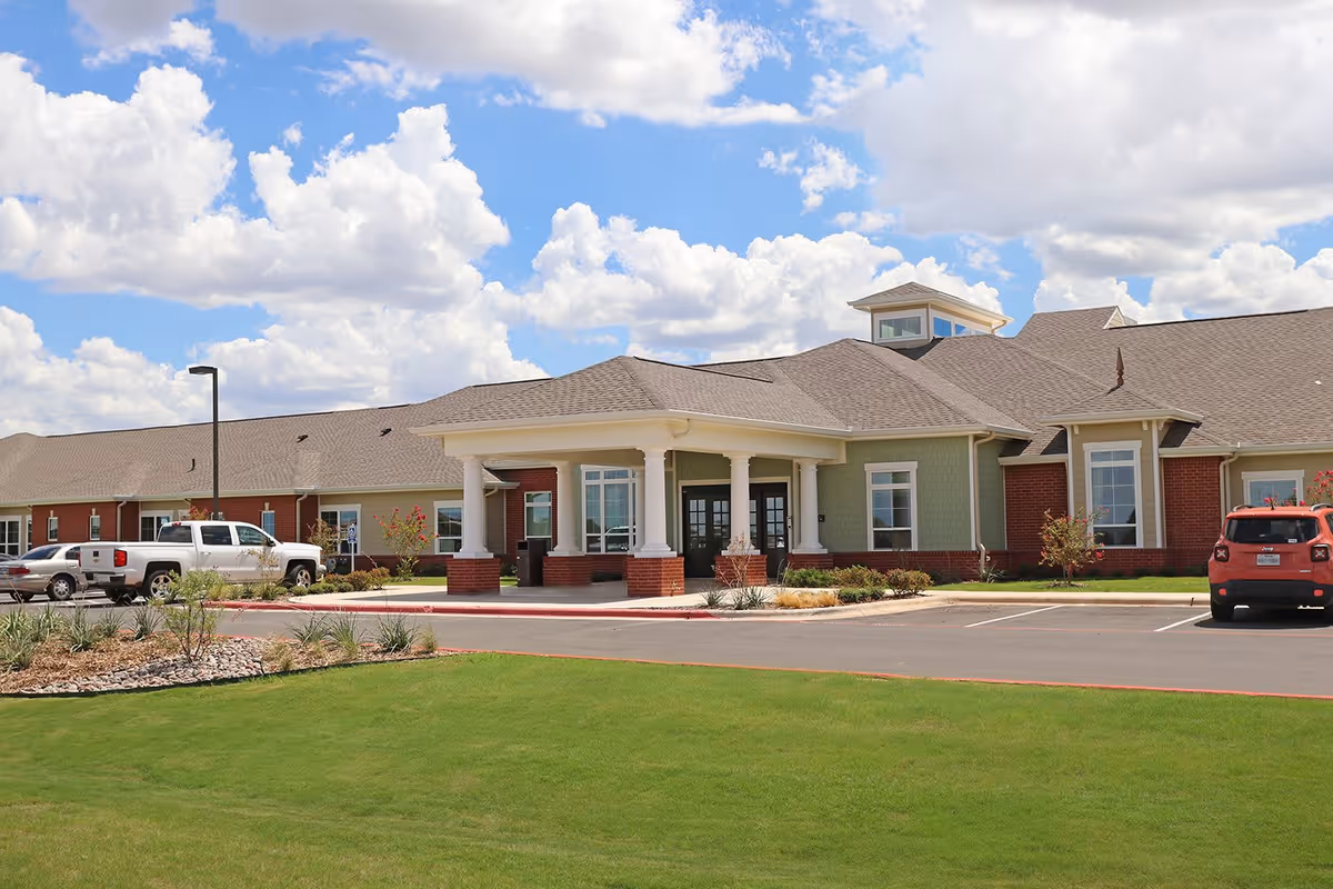 Exterior view of a single-story senior living facility building with a covered entrance supported by white columns, surrounded by a well-maintained lawn and parking spaces with several vehicles parked. The sky is partly cloudy with blue patches visible.