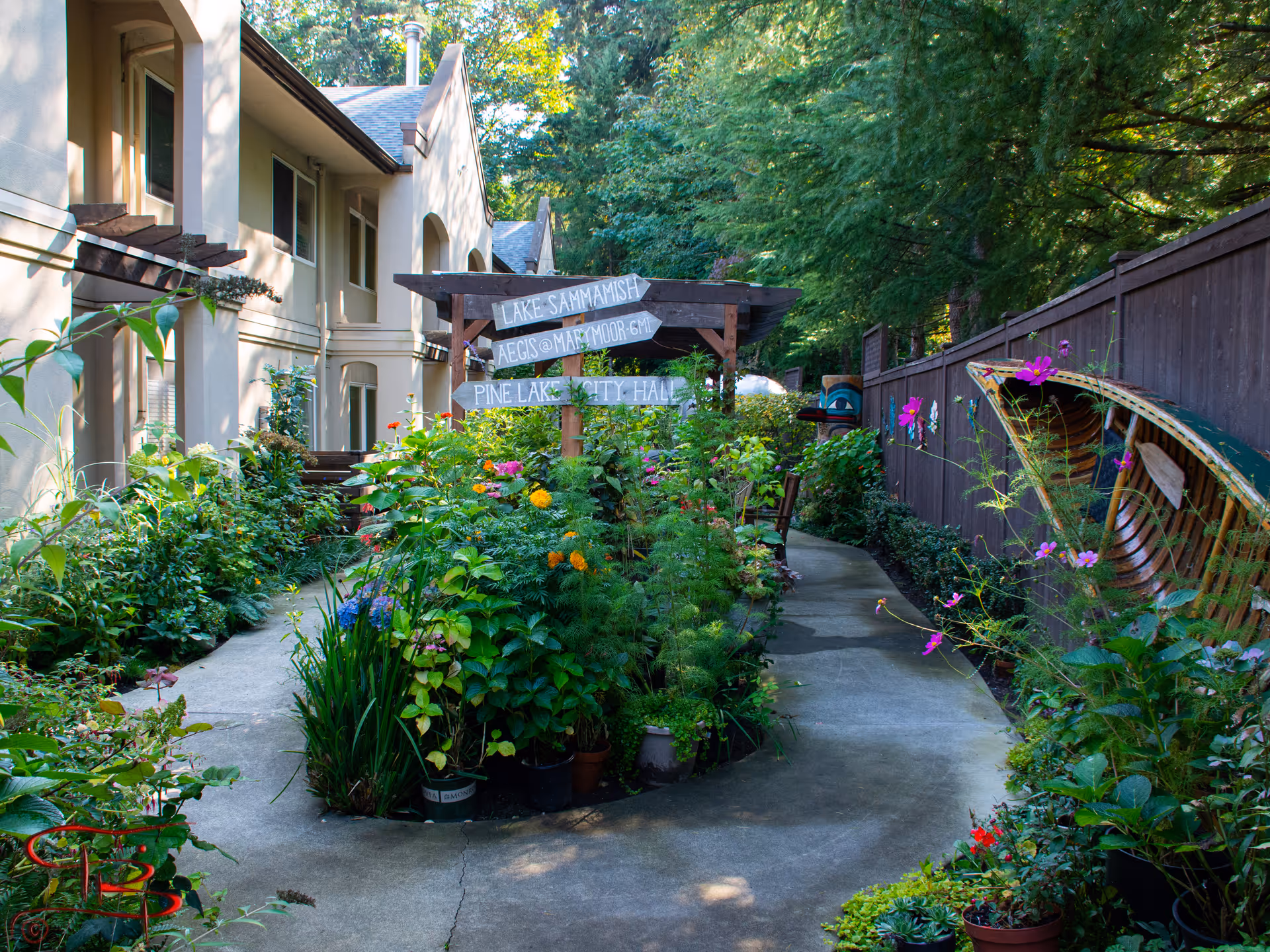 A serene outdoor garden area at Aegis Living Marymoor with a winding concrete pathway surrounded by lush green plants and colorful flowers. A wooden pergola with directional signs points to Lake Sammamish, Aegis @ Marymoor, and Pine Lake City Hall. The garden is bordered by a wooden fence on one side and the building on the other, with trees providing shade.