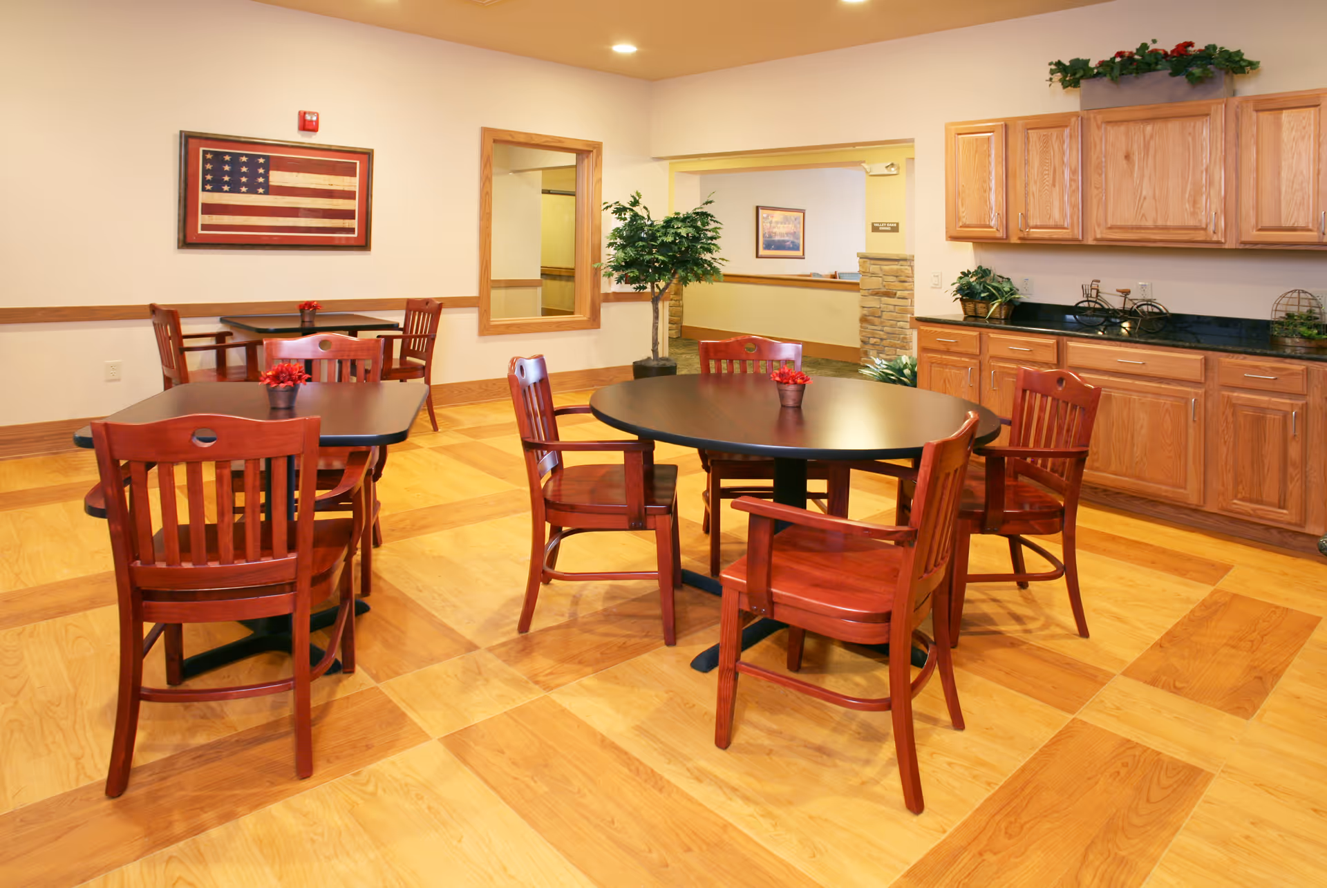 A dining area with wooden floors and several dark round and square tables surrounded by wooden chairs. There are small red flower arrangements on each table. On the right side, there are wooden cabinets with a black countertop and some decorative plants. A framed American flag hangs on the wall, and a potted plant is placed near an open doorway leading to another room.