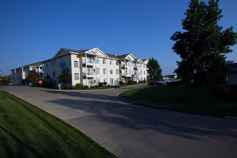 Exterior view of a three-story senior living facility building with balconies, surrounded by green lawns and trees under a clear blue sky.