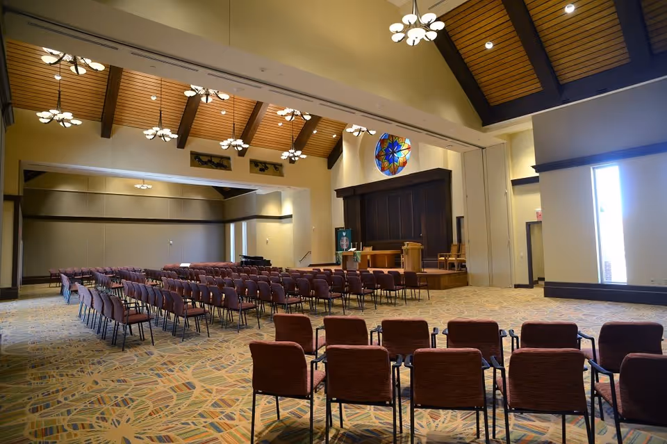 Interior view of a large meeting or chapel room with rows of brown chairs arranged facing a wooden stage with a pulpit and chairs. The room has a high ceiling with wooden beams, multiple hanging light fixtures, a colorful stained glass window above the stage, and patterned carpet flooring.
