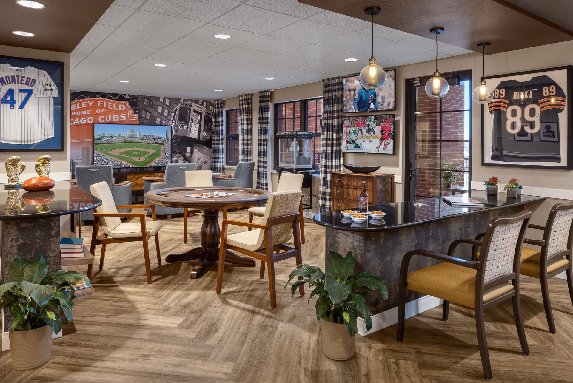 A cozy senior living common area with a round wooden game table surrounded by chairs, a curved countertop with snacks and drinks, framed sports jerseys and photos on the walls, large windows with plaid curtains, and plants adding greenery to the space.
