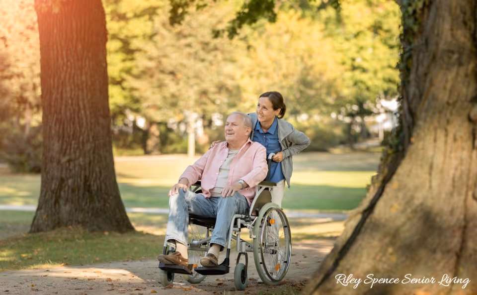 An elderly man sitting in a wheelchair being pushed by a woman along a tree-lined path in a park or garden area during daytime with sunlight filtering through the trees.