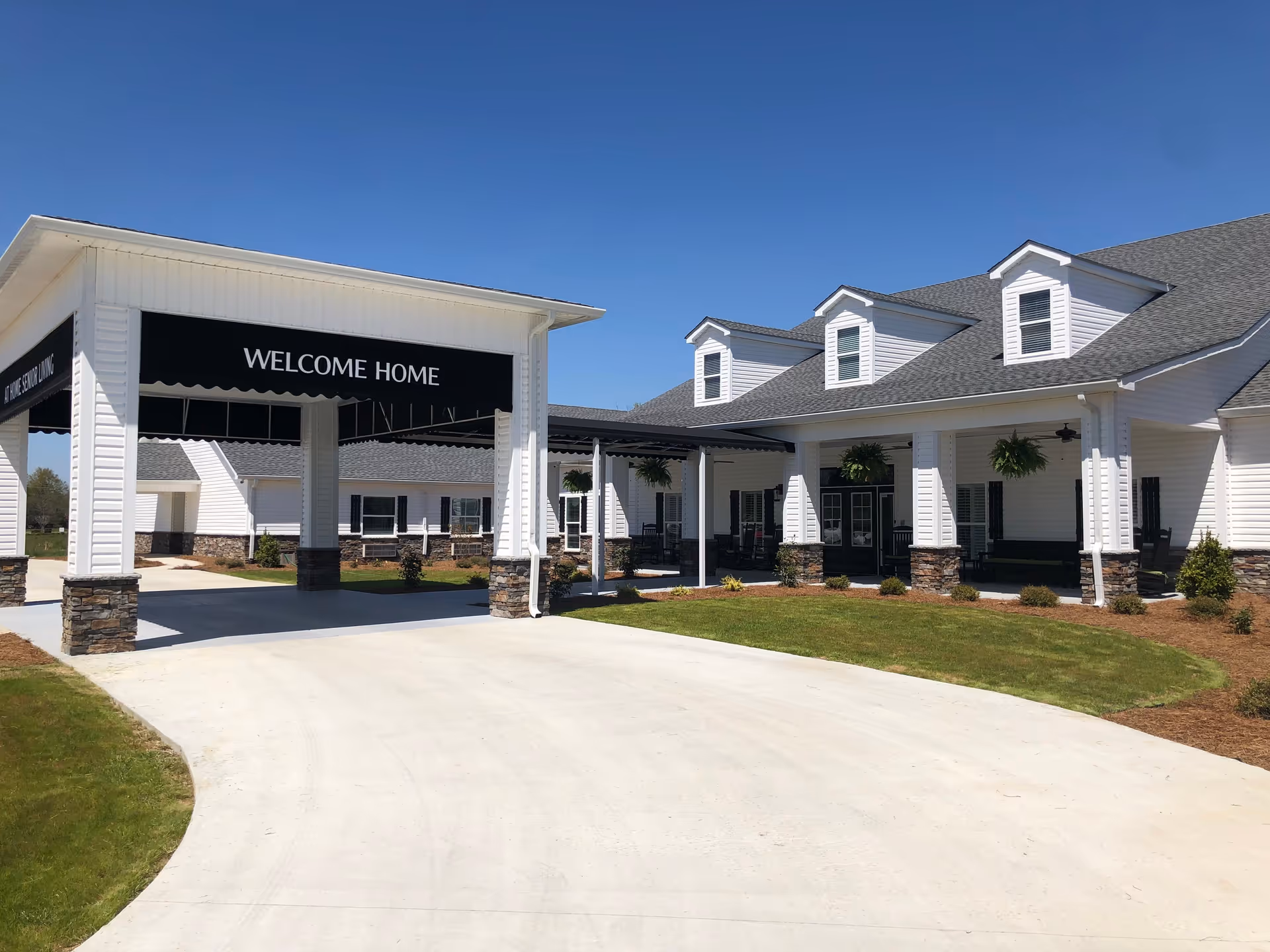 Exterior view of a senior living facility building with white siding and stone accents. The entrance features a covered driveway with a black awning that reads 'WELCOME HOME'. The building has multiple dormer windows on the roof and hanging plants on the porch. The sky is clear and blue.