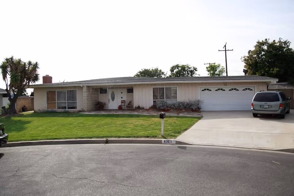 Single-story residential house with a front lawn, a driveway with a parked minivan, a mailbox, and a garage with two doors. The house has a white exterior with large windows and a front door with an oval glass panel.