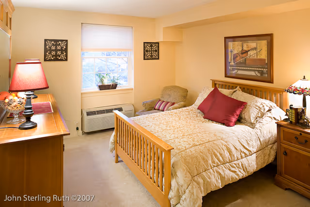 A cozy bedroom with a wooden bed frame, beige patterned bedding, and two red accent pillows. There is a wooden nightstand with a decorative lamp and books on the right side of the bed. A framed picture hangs on the wall above the bed. To the left, there is a wooden dresser with a red lamp and a bowl of fruit. A window with a white blind and a small plant on the windowsill is visible, along with a beige armchair with a striped pillow in the corner.