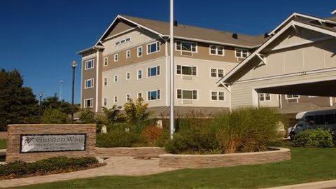 Exterior view of a multi-story beige and brown retirement community building with landscaped greenery and a stone sign that reads 'Garden Way Retirement Community' in front under a clear blue sky.