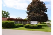 Exterior view of a single-story building with a flat roof, surrounded by green bushes and a large tree. A sign in front reads 'Cortland Park Rehabilitation And Nursing Center'. The sky is partly cloudy.