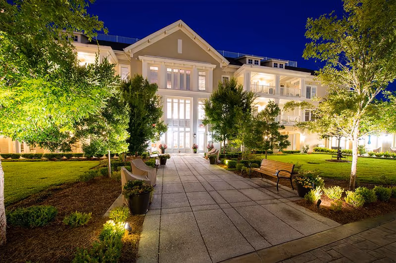 Night view of the entrance to a large, well-lit senior living facility building with a paved walkway lined by trees, benches, and plants on both sides.