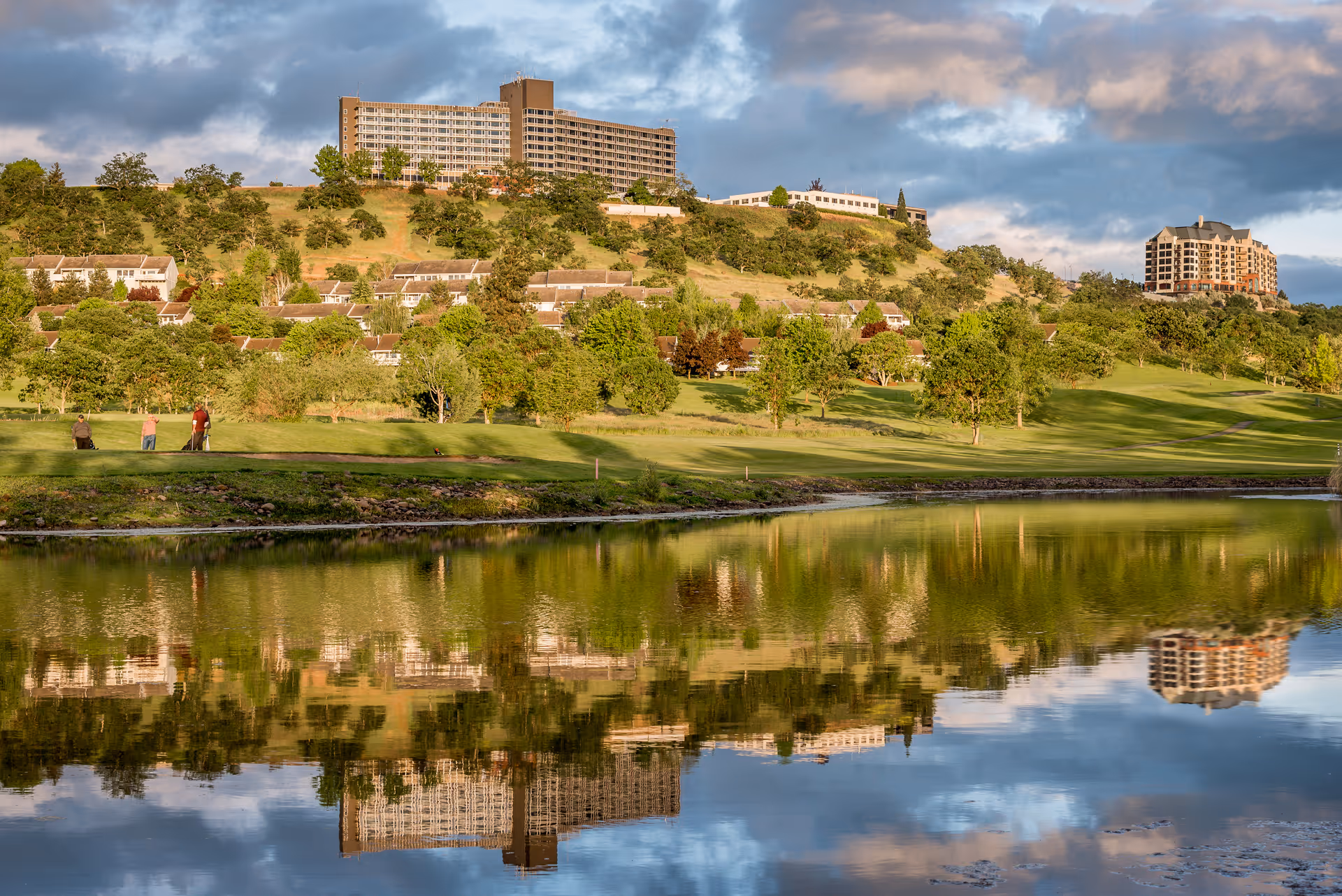 A scenic view of a hillside with multiple buildings, including a large multi-story building at the top and smaller residential buildings below. The hillside is covered with green trees and grass, and there is a calm body of water in the foreground reflecting the buildings and sky. Several people are seen standing on the grassy area near the water.
