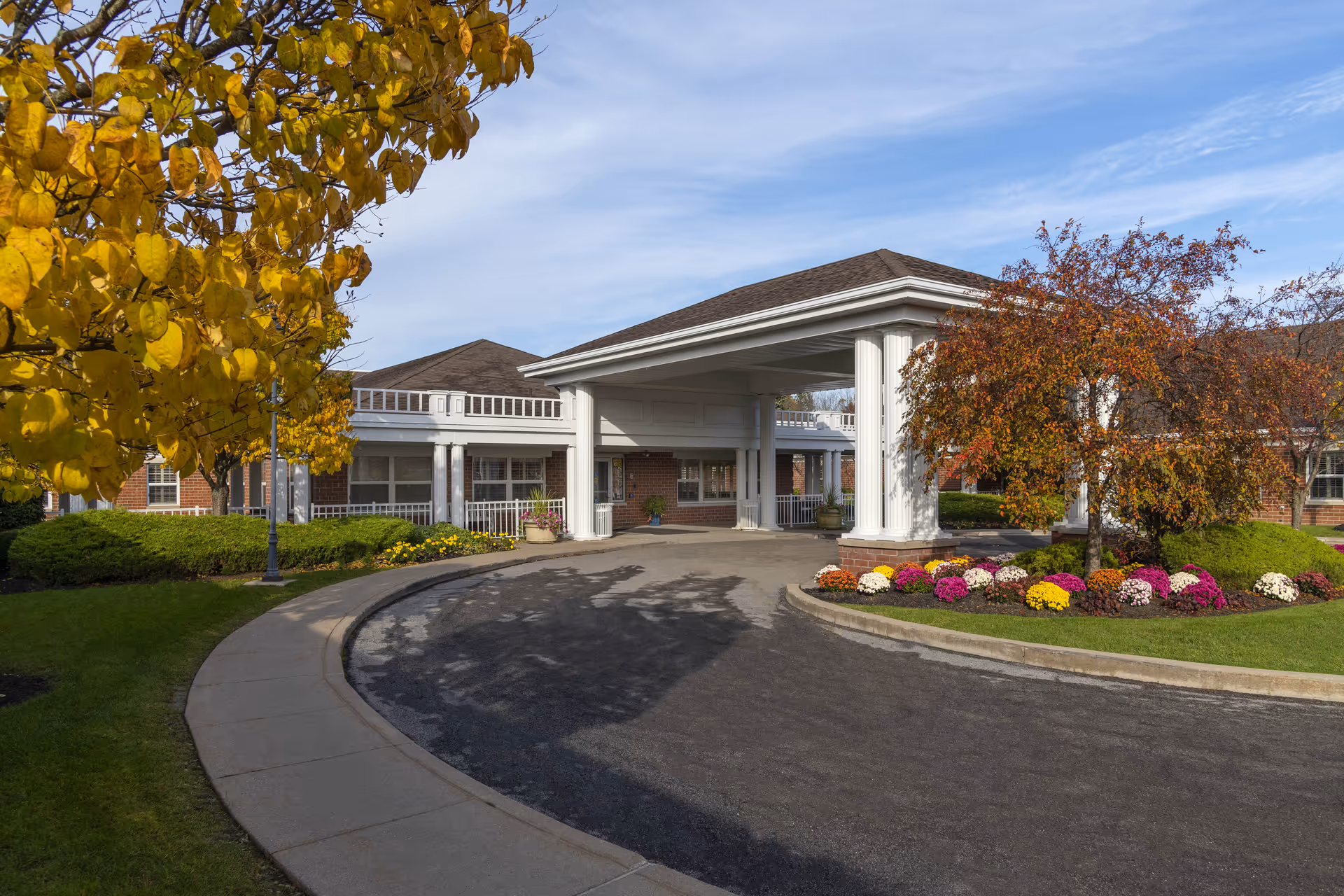 Exterior view of a senior living facility entrance with a covered driveway supported by white columns. The building has a brick facade and a brown roof. There are landscaped areas with green bushes, colorful flowers, and trees with autumn foliage surrounding the driveway.