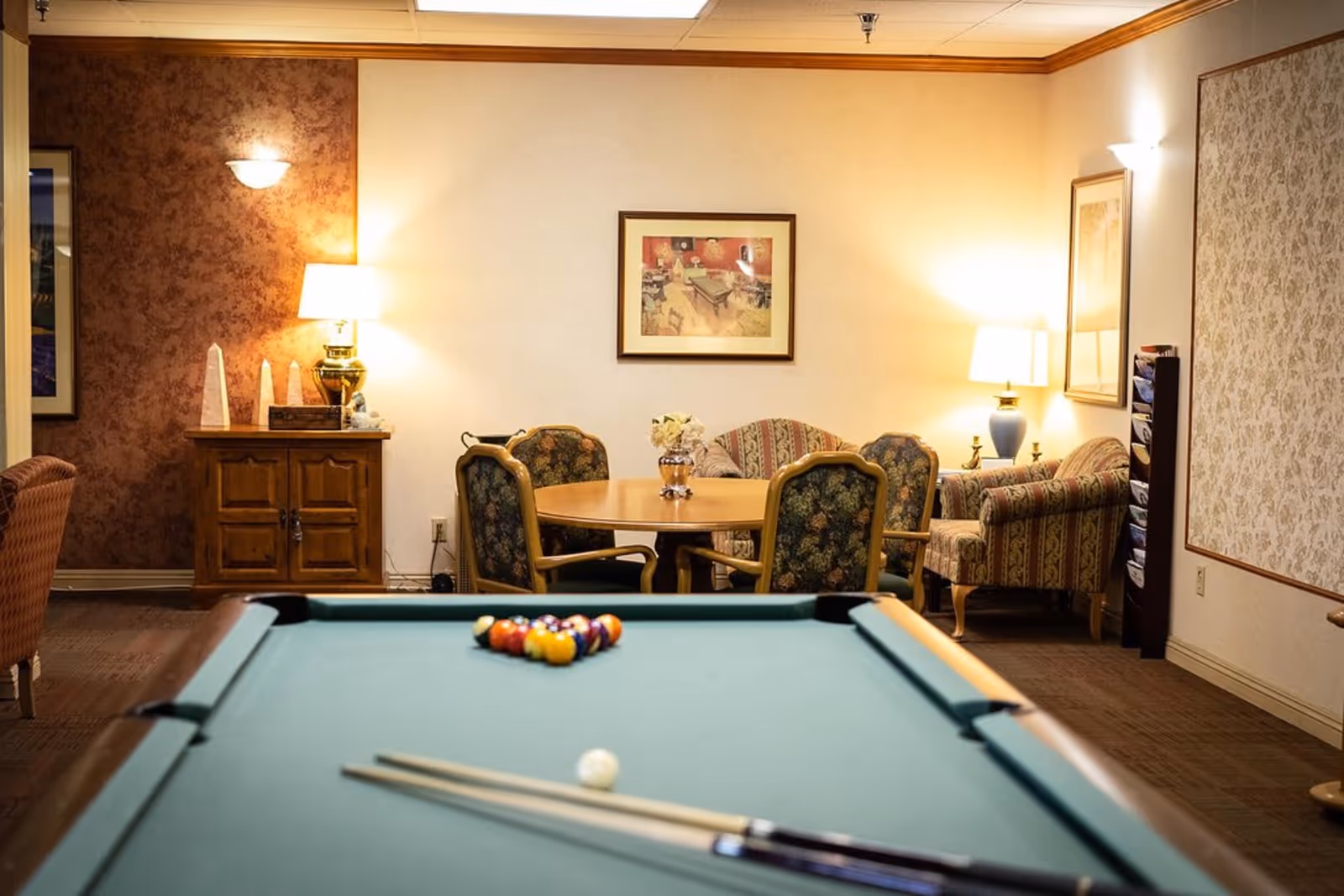 A cozy recreational room featuring a pool table in the foreground with billiard balls and cues arranged on it. In the background, there is a round wooden table surrounded by four upholstered chairs with floral patterns. The room is warmly lit by table lamps on wooden furniture and has framed artwork on the walls, along with patterned wallpaper and carpeted flooring.