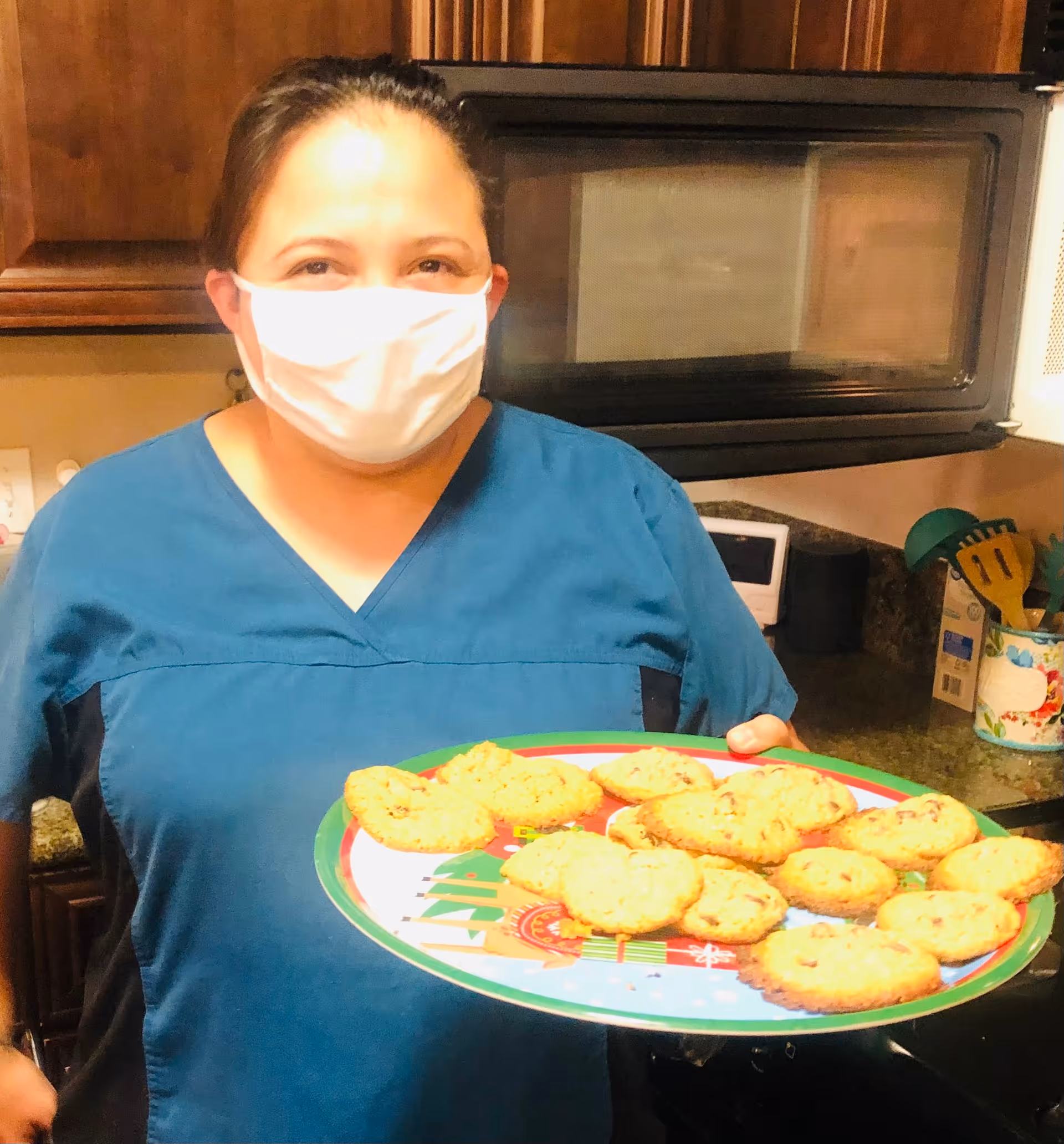 A woman wearing a blue scrub top and a white face mask is standing in a kitchen holding a festive plate with freshly baked cookies. Behind her, there are wooden cabinets, a microwave, and kitchen utensils on the counter.