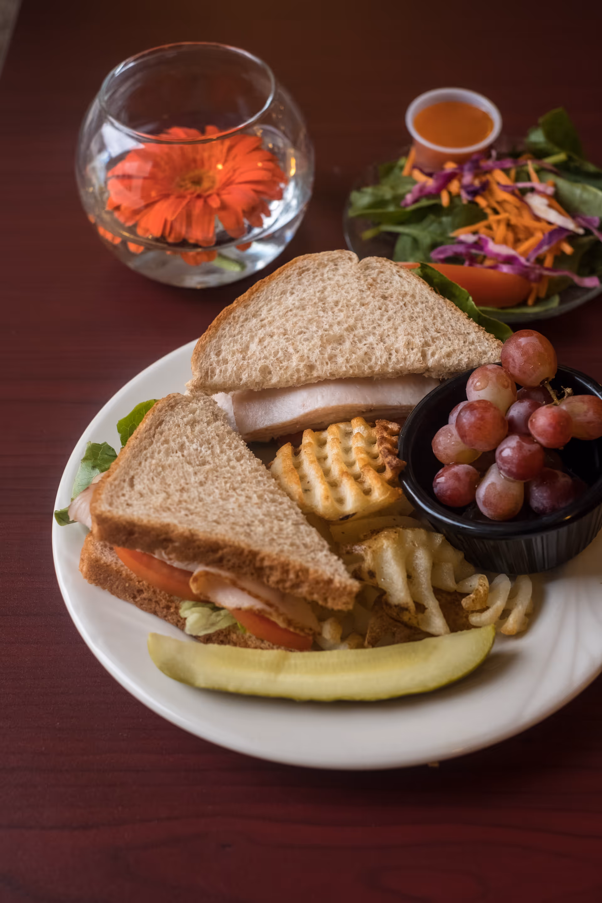 A plate with a sandwich made of whole wheat bread, lettuce, tomato, and turkey, served with waffle fries, a pickle spear, and a small cup of red grapes. In the background, there is a small bowl of salad with shredded carrots, purple cabbage, and a cup of dressing, along with a glass bowl containing an orange flower in water on a wooden table.