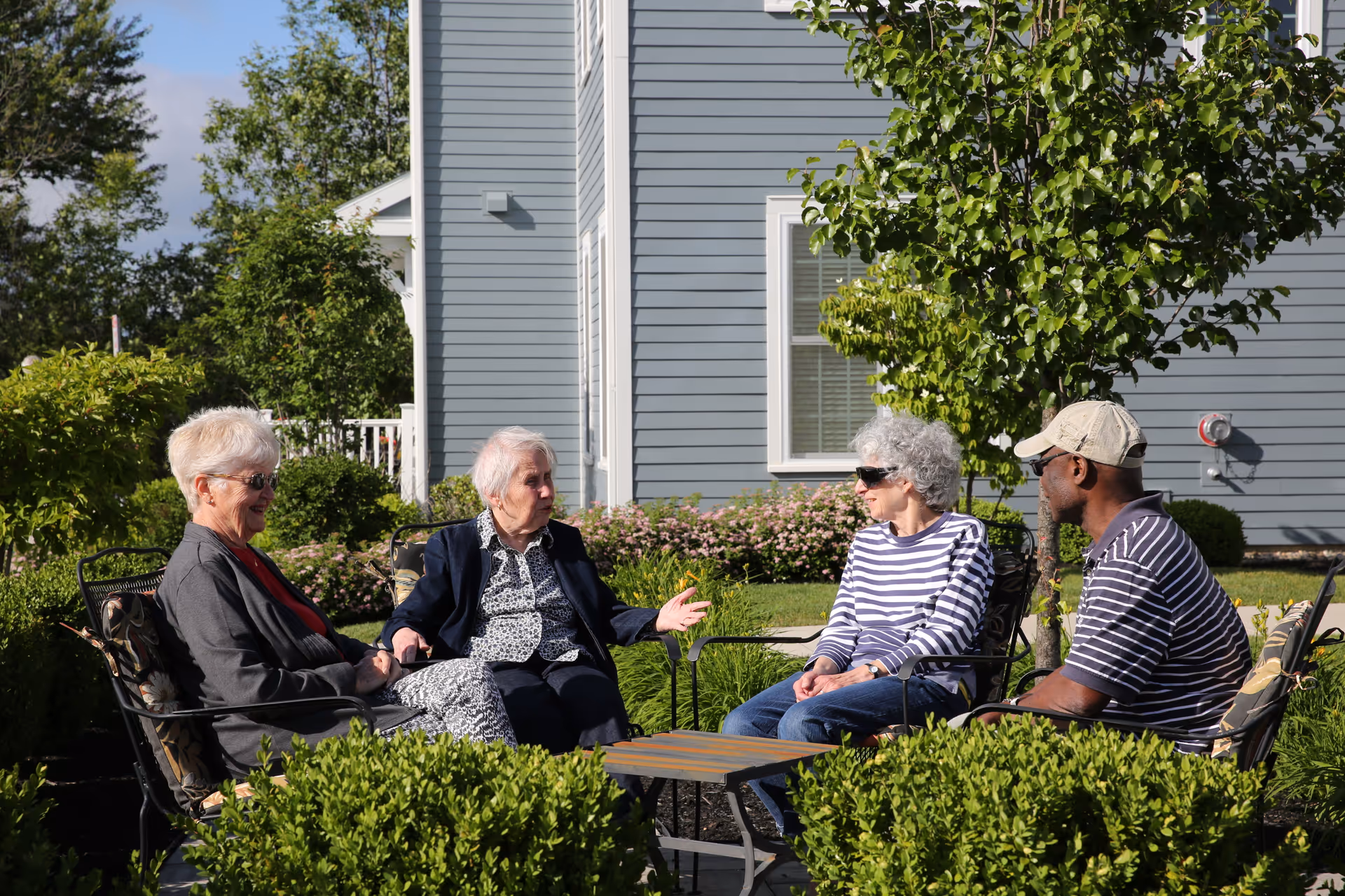 Four elderly people sitting outdoors in a garden area, engaged in conversation. They are seated on metal chairs around a small table, with green bushes and a tree nearby. The background shows the exterior of a light blue building with white trim and windows.