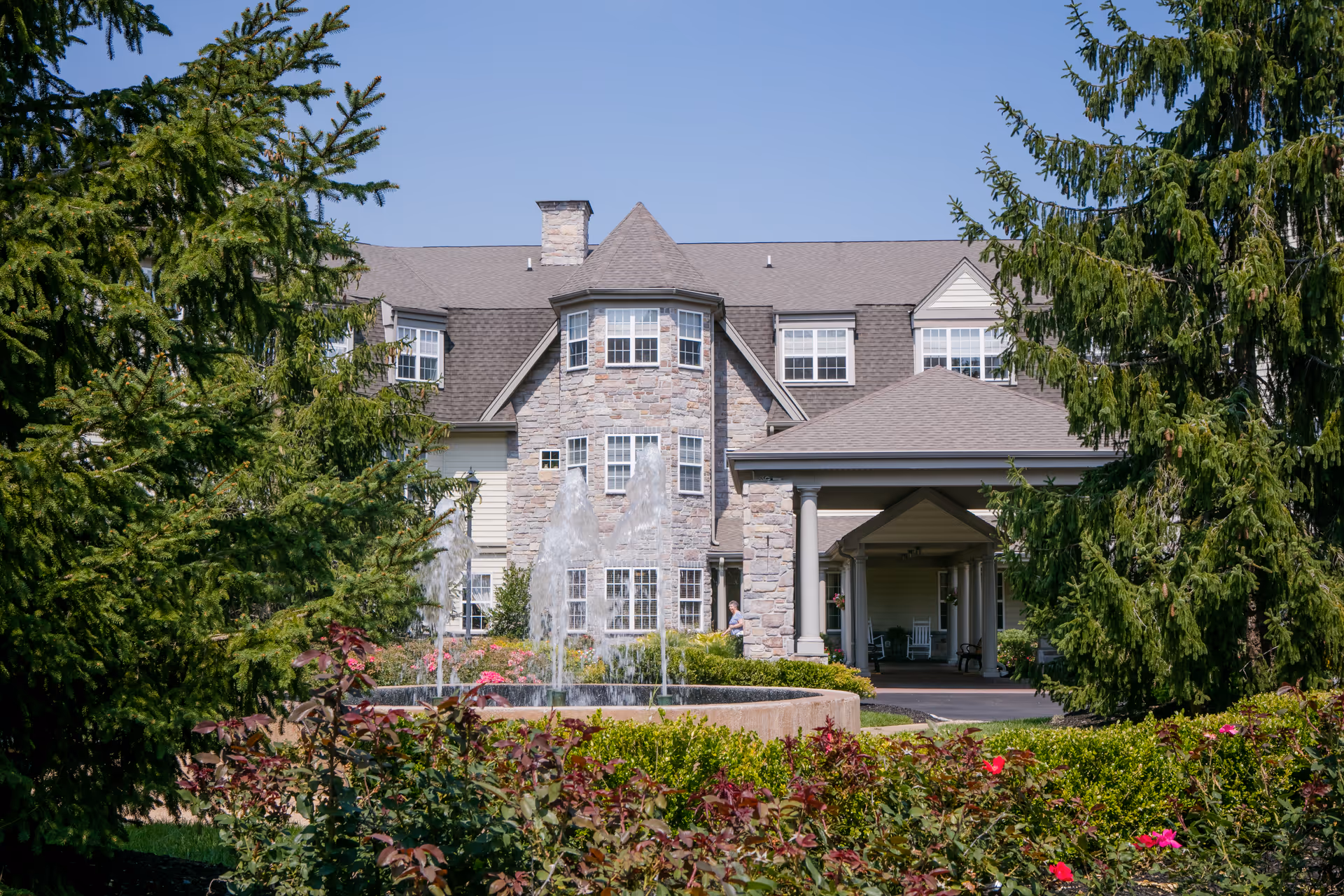 Exterior view of a senior living facility building with stone and siding facade, a covered entrance supported by columns, surrounded by green trees, bushes, and a water fountain in front.