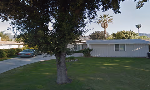 Single-story residential building with a gray exterior, a large tree in the front yard, a green lawn, and a driveway with a parked car. Palm trees and mountains are visible in the background under a clear sky.