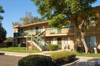 Exterior view of a two-story senior living facility building with beige walls and green railings. There is a central staircase leading to the upper floor, surrounded by well-maintained bushes, trees, and a grassy area under a clear blue sky.