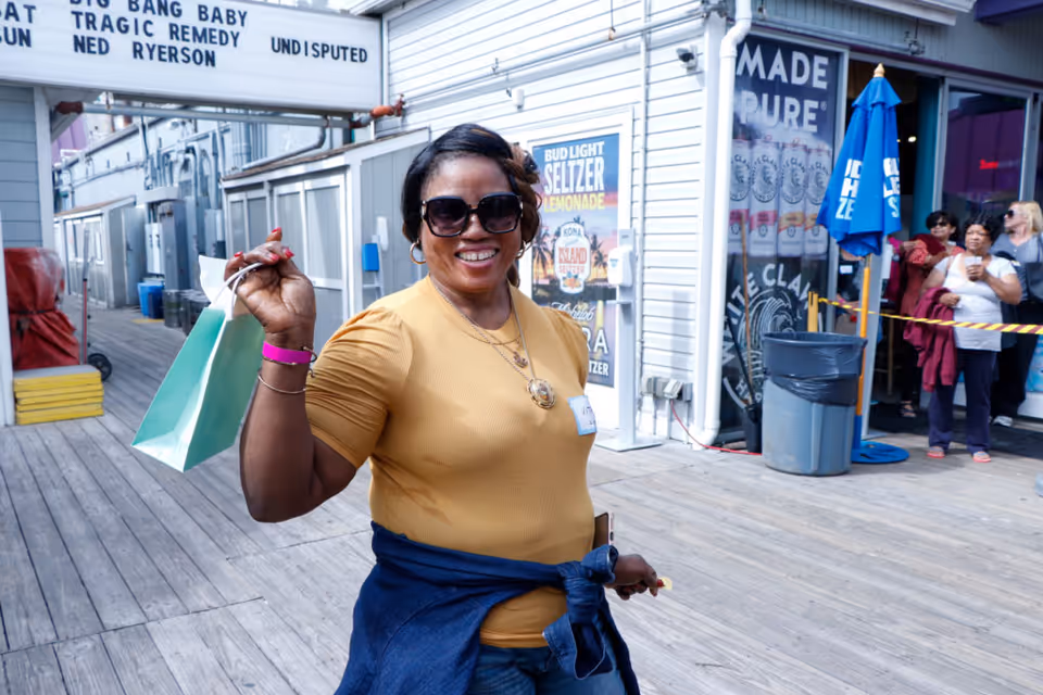 A smiling woman wearing sunglasses and a yellow shirt holds up a small shopping bag while standing on a wooden boardwalk outside a building with signs and other people in the background.