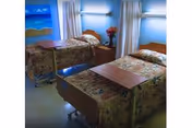 A nursing facility room with two hospital beds covered in floral bedspreads. Each bed has an overbed table, and there is a nightstand with a vase of flowers between the beds. The room has light blue walls, curtains, and a wall-mounted light fixture above each bed. A mural of a beach scene is visible on one wall.