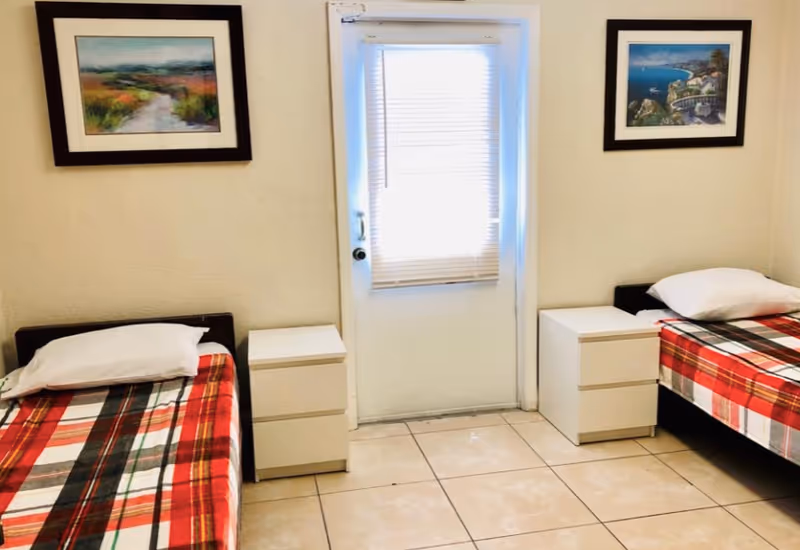A small bedroom with two single beds on either side of a door with a window covered by blinds. Each bed has a white pillow and a red, black, and white plaid blanket. Between each bed and the door is a white nightstand with two drawers. Two framed landscape paintings hang on the walls above each bed. The floor is tiled with light-colored tiles.