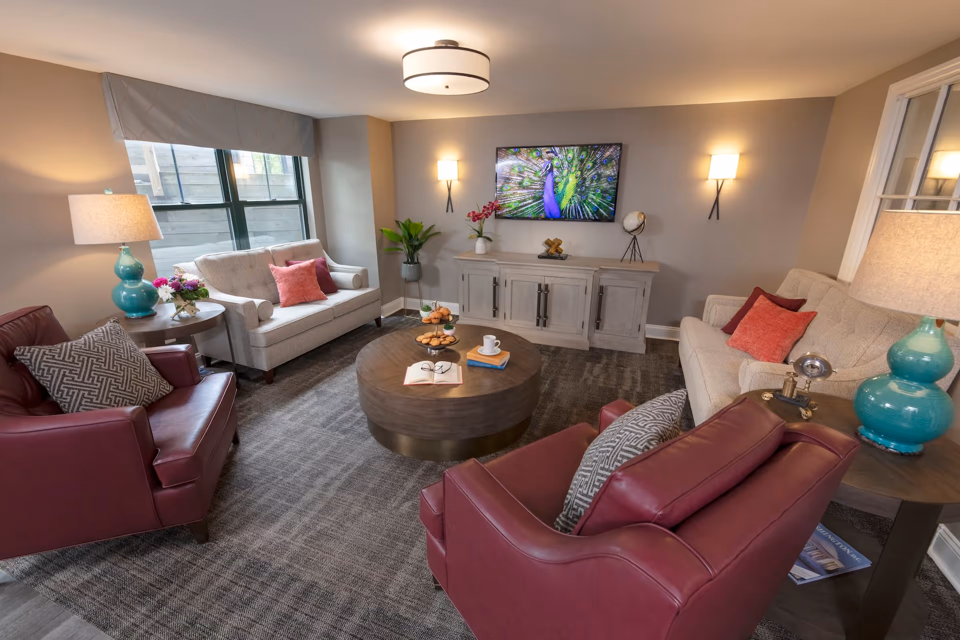 A cozy living room with two beige sofas and two red leather armchairs arranged around a round wooden coffee table. The coffee table has an open book, a pair of glasses, a cup, and a tiered tray with muffins. There are two turquoise lamps on side tables, a large window with a gray valance, a flat-screen TV mounted on the wall displaying a peacock, and decorative plants and items on a wooden cabinet beneath the TV. The room has beige walls and a gray carpet.