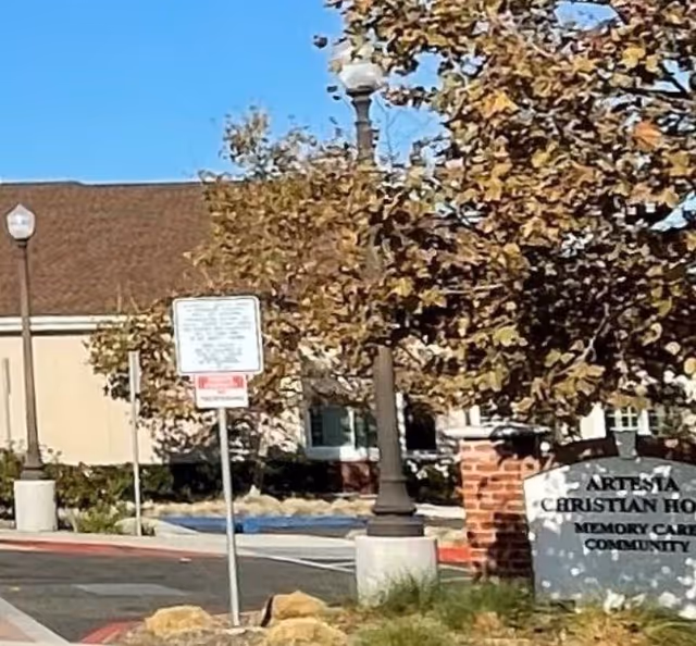Outdoor view of Artesia Christian Home showing part of the building, a tree with autumn leaves, street lamps, a parking area with a handicap parking space, and a sign indicating the facility name and that it is a memory care community.