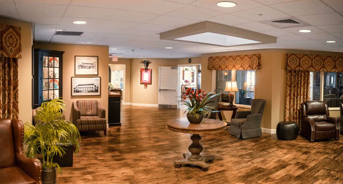 A spacious and warmly decorated common area with wooden flooring, comfortable armchairs, a round wooden table with a floral arrangement, framed pictures on the wall, and large windows with patterned valances and curtains.